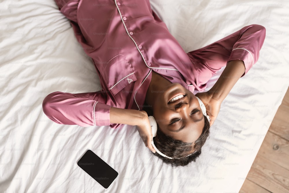 Application musicale. Femme noire du millénaire écoutant de la musique en ligne dans des écouteurs à l’aide d’un téléphone allongée dans son lit et se relaxant les yeux fermés à la maison, au-dessus de la vue. Excellent concept de playlist