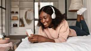 Happy African American Lady Using Phone Wearing Headphones Lying In Bed At Home. Black Millennial Woman Using Mobile Application Browsing Internet Via Smartphone. New App For Cellphone. Panorama