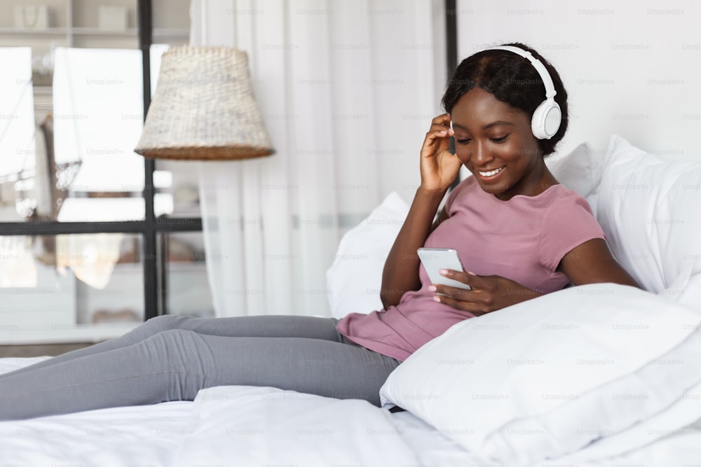 Cheerful Black Female Using Smartphone Listening To Music Wearing Headphones Sitting In Bed At Home. Student Lady Listens Podcast. Musical Mobile Application, Technology And Fun Concept