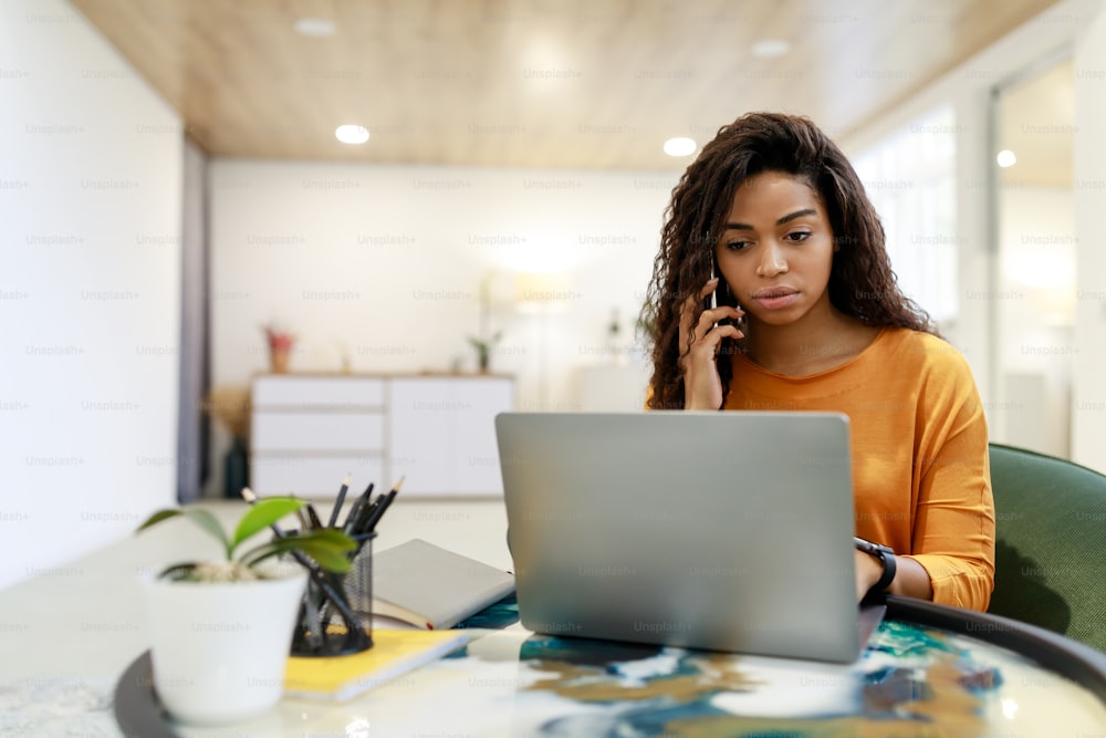 Mobile Communication. Portrait Of Focused Black Female Freelancer Talking On Cellphone Working On Pc At Home Office, Lady With Distance Job Using Computer Having Phone Conversation, Free Copy Space