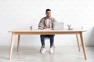 Gérant masculin souriant du Moyen-Orient travaillant sur un ordinateur portable dans un bureau moderne, un gars joyeux assis au bureau et utilisant un PC, regardant la saisie d’écran sur le clavier, appréciant le travail à distance, toute la longueur du corps