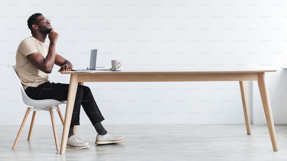 Side view of pensive young black guy using laptop, sitting at desk, thinking over something, working online from home office, free space. Dreamy African American man having remote job