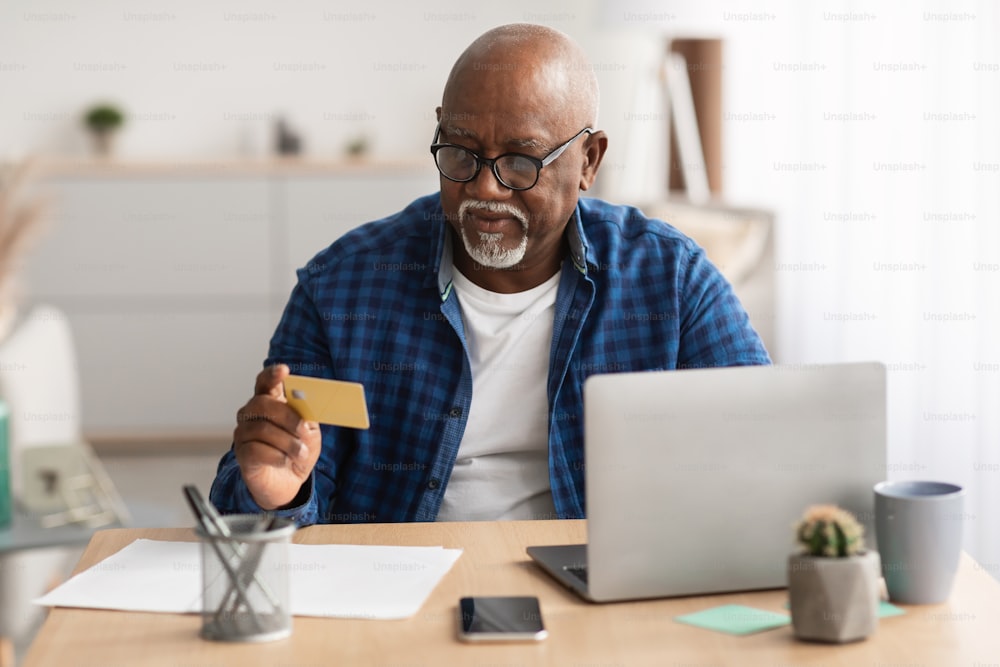 Senior African American Businessman Shopping Online Using Credit Card And Laptop Making Payment Purchasing Something Sitting At Workpalce Indoor, Wearing Glasses. Ecommerce Concept