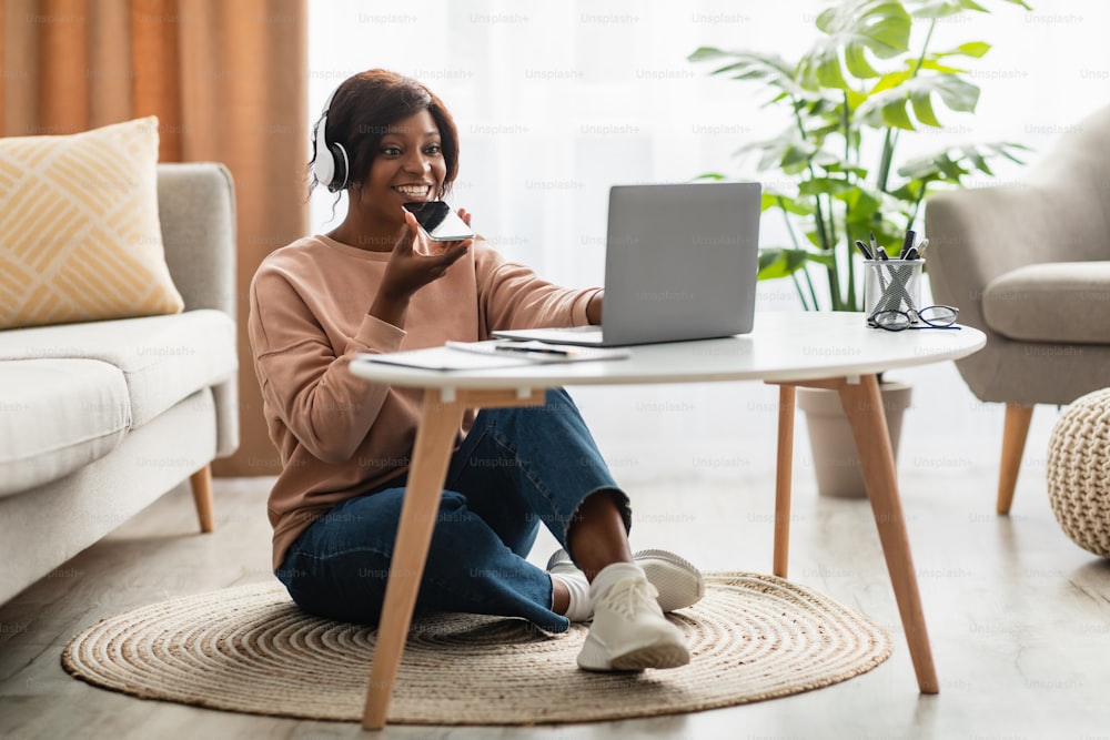 흑인 밀레니얼 여성은 스마트폰에서 음성 검색 애플리케이션을 사용하여 노트북 컴퓨터에 앉아 집에서 무선 헤드폰을 착용하고 있다. 가제트 라이프 스타일과 현대 통신 개념