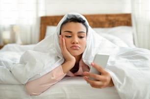 Upset young black woman looking at smartphone screen while laying in bed under blanket, feeling lonely or depressed. Sad african american lady waiting for call from lover