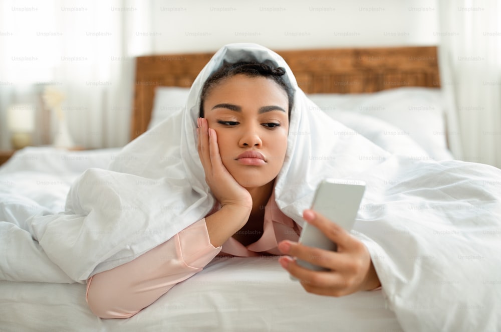 Upset young black woman looking at smartphone screen while laying in bed under blanket, feeling lonely or depressed. Sad african american lady waiting for call from lover