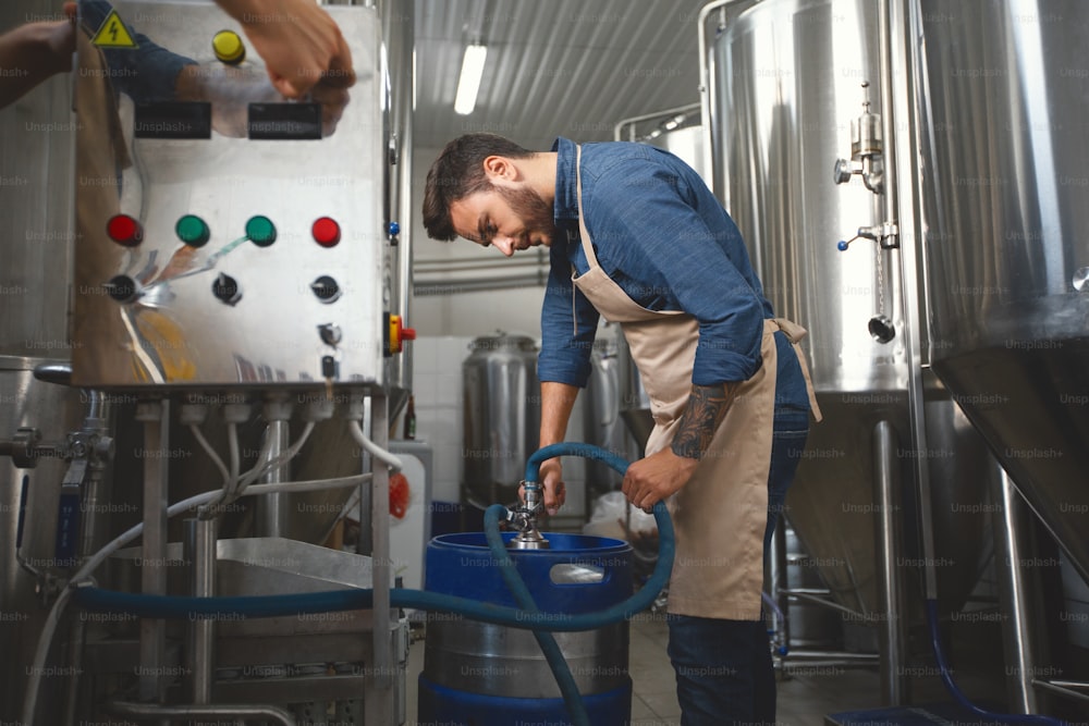 Equipment in modern factory, brewery, employee work with craft beer. Busy contented millennial handsome bearded guy in apron, filling bottles with alcohol drink in plant, on metal kettles background