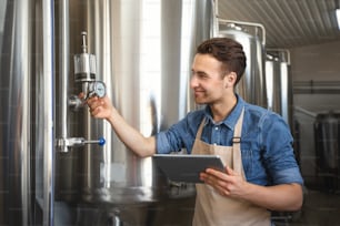 Worker, brewer, business owner works on factory with equipment, modern tech. Satisfied smiling young attractive guy in an apron holds tablet and checks metal boilers at plant in brewery interior