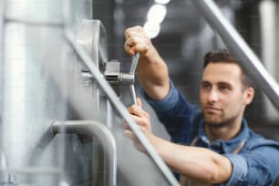 Factory work, equipment management, brewing and own business. Portrait of concentrated young attractive man turns valve on metal boiler at plant, selective focus, blurred, close up, empty space