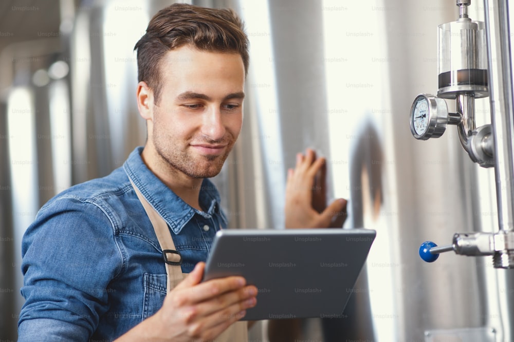 Modern business, app for plant management, own brewery, alcohol drink production. Portrait of glad millennial handsome male in apron looks at tablet and checks metal boilers at factory, empty space