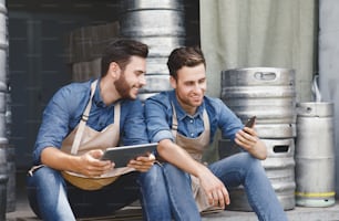Brewery, tech, online ordering and sales of products, successful family business. Rest and break. Happy attractive young men in aprons sit in warehouse with metal kegs and look at phone and tablet