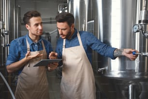 Successful modern business, work in alcohol drink factory, professionals with gadget at craft brewery. Busy millennial attractive men workers in aprons look at tablet and check metal boilers at plant