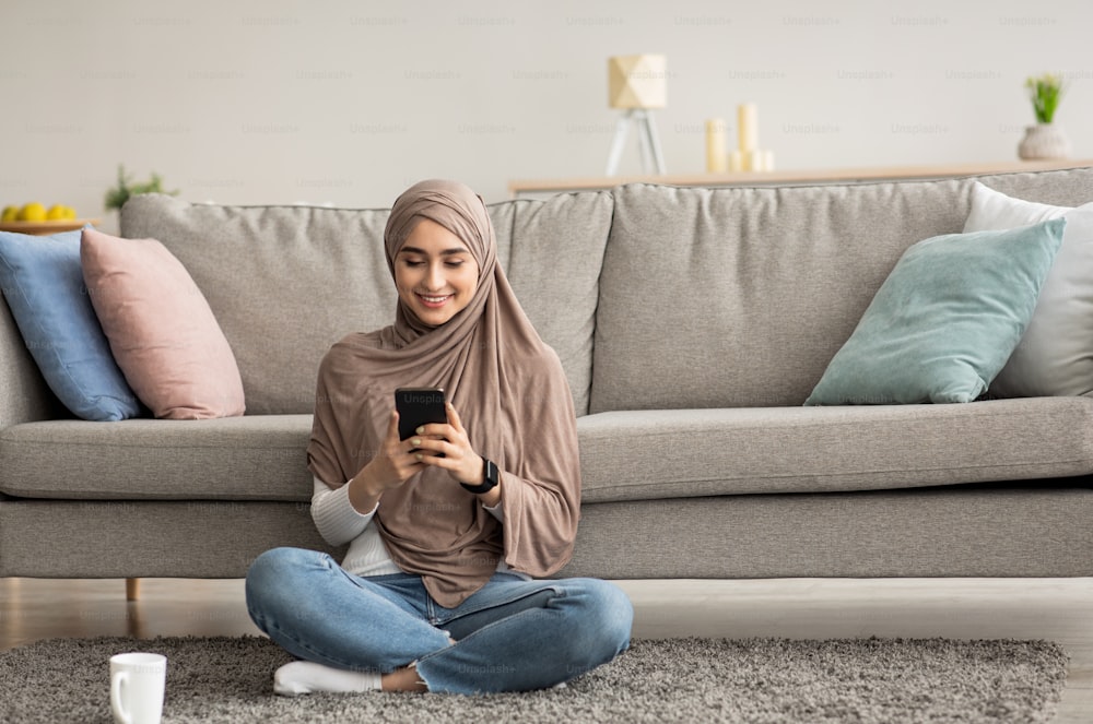 Modern relaxation at home with gadget, coffee break and chat in social networks. Smiling millennial arab lady in hijab typing message on smartphone sits on floor with cup of drink, in living room
