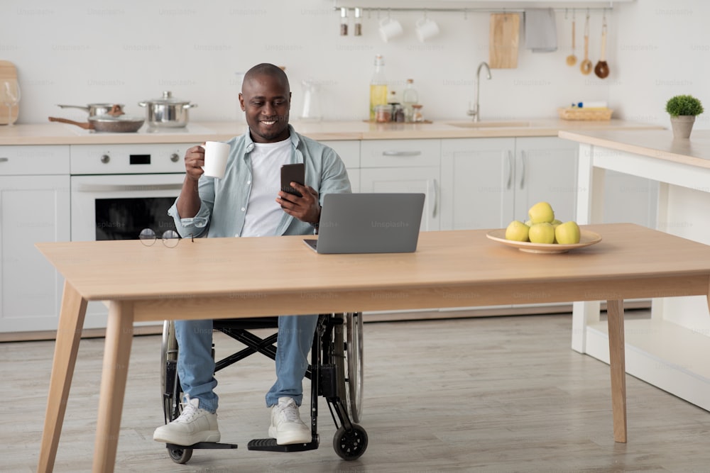 Morning coffee, news, chat in social networks, work and education at home. Smiling adult african american male disabled in wheelchair drinking, look at phone, at table with laptop in kitchen interior