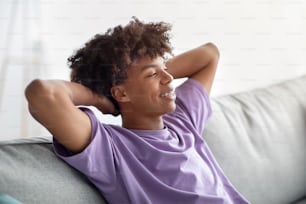 Carefree black teen guy relaxing on couch with hands behind his head, spending time at home during covid lockdown. Peaceful African American youth resting on sofa, having lazy weekend