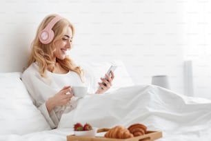 Healthy breakfast alone, surfing in internet, watch video in gadget. Smiling cute millennial european lady in headphones sitting on comfortable bed in white bedroom with food and typing on smartphone