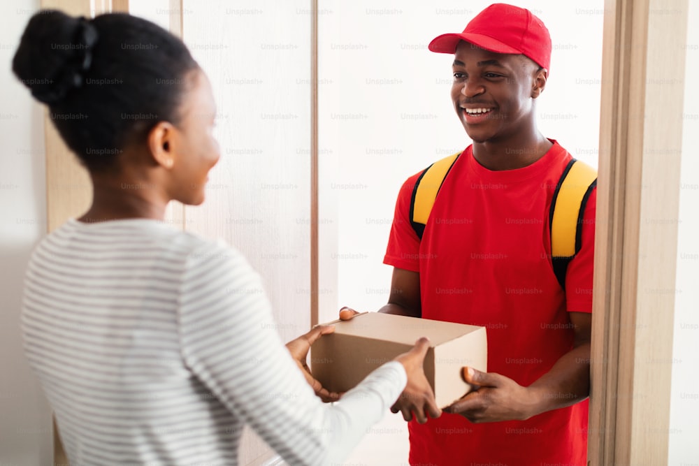 쾌활한 흑인 택배 남자가 실내에서 문 앞에 서 있는 여자에게 골판지 소포 상자를 배달한다. 빠른 배송 서비스 및 우편 배송 개념. 선택적 초점