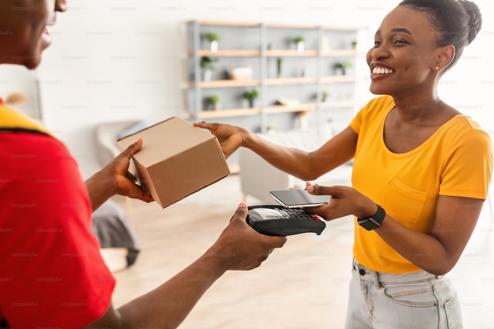 African American Customer Woman Paying Delivery Guy Using Phone Receiving Parcel Box And Putting Smartphone To Payment Terminal Indoors. Courier Service Concept. Cropped, Selective Focus