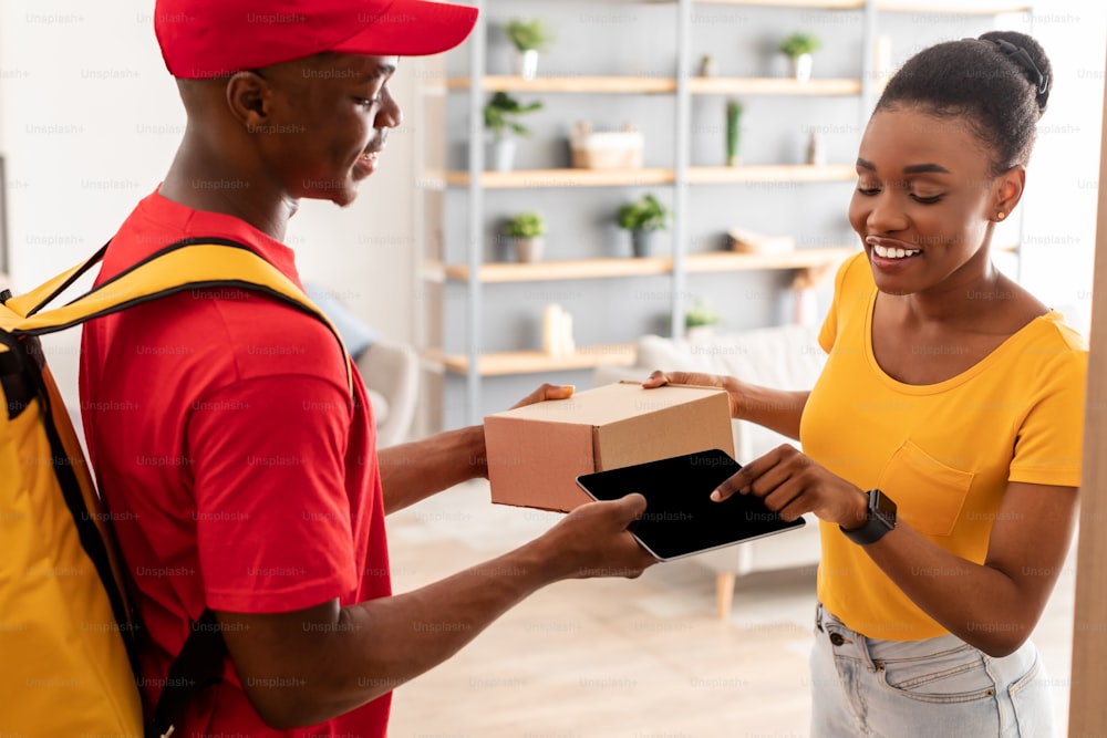 Black Lady Receiving Parcel From Courier And Scanning Fingerprint For Personal Verification And Smart Payment Via Digital Tablet Standing At Doors Of Her Home. Innovative Delivery Service