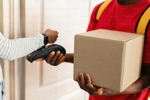 Cropped Shot Of Customer Lady Paying Courier For Parcel Delivery Using Smartwatch, Putting Watch To Payment Terminal At Home. Selective Focus On Cardboard Box