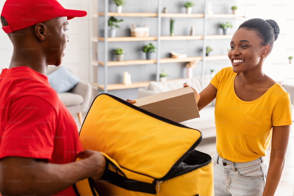 택배 서비스 개념입니다. 빨간 유니폼을 입고 집 출입구에 서 있는 문에 주문을 배달하는 흑인 여성 고객에게 상자를 주는 배달원. 선택적 초점