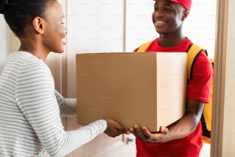 アフリカ系アメリカ人の女性の顧客に小包を配達する制服を着たプロの宅配便業者は、彼女の家の戸口に立っている板紙箱を与えます。パッケージ配信のコンセプト。トリミングされた、選択フォーカス