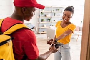 Smart Payment. African American Lady Paying Courier Guy With Smartwatch Banking Application Receiving Parcel At Home. E-Commerce, People And Gadgets Concept. Selective Focus