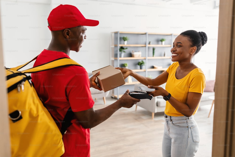 Door Delivery. Happy Black Customer Lady Receiving And Paying For Package Box With Phone Taking Parcel From Courier Guy In Red Uniform Standing In Doorway At Home. Modern Shopping Concept