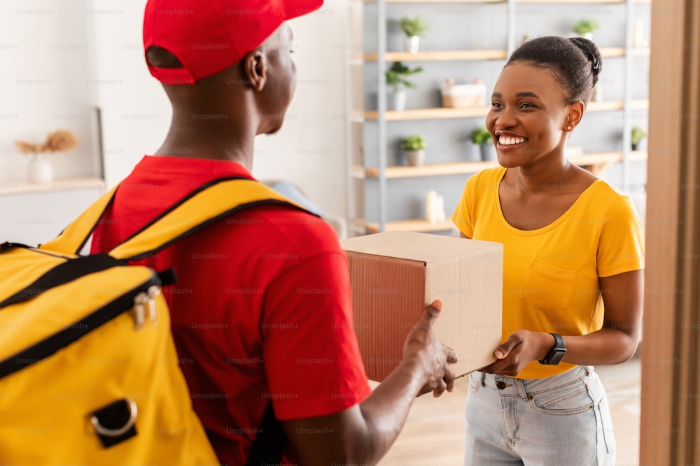 Shopping Delivery. Black Male Courier Guy Giving Big Box To Joyful Female Customer Standing In Doors Of Her Home. Man In Uniform Delivering Parcel To African American Lady. Selective Focus