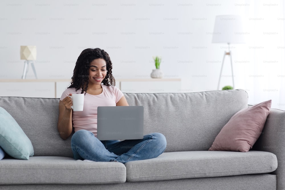 Businesswoman working on computer sit at home, hold coffee cup in hand, managing business, freelancing. Smiling millennial african american woman typing on laptop and drinks in living room interior