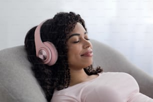 Peace of mind, listen favorite music, enjoy modern technology at home at spare time. Satisfied happy young african american lady in wireless headphones rest, relax in living room interior, close up