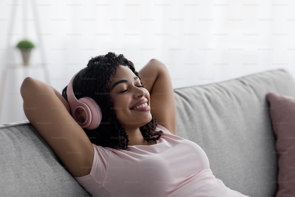 Relax with favorite music, rest and break with mobile gadget. Happy satisfied millennial african american female in wireless headphones resting on sofa with closed eyes in minimal room interior
