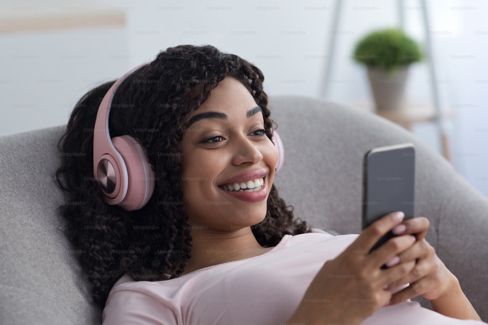 Watch funny video, new app, chat on social networks at home. Satisfied glad cute millennial african american female on wireless headphones on sofa, enjoying comfort with smartphone or listen music