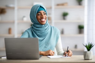 Pensive Black Muslim Businesswoman In Hijab Sitting At Desk With Laptop Computer In Office And Looking Away, Smiling African Islamic Lady Thinking About Future Plans And Taking Notes To Notepad