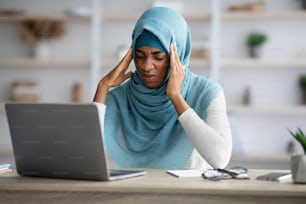 Concepto de dolor de cabeza. Mujer independiente musulmana negra estresada que sufre de migraña en el lugar de trabajo, joven mujer islámica africana molesta sentada en el escritorio con computadora portátil y tocando sienes, espacio libre