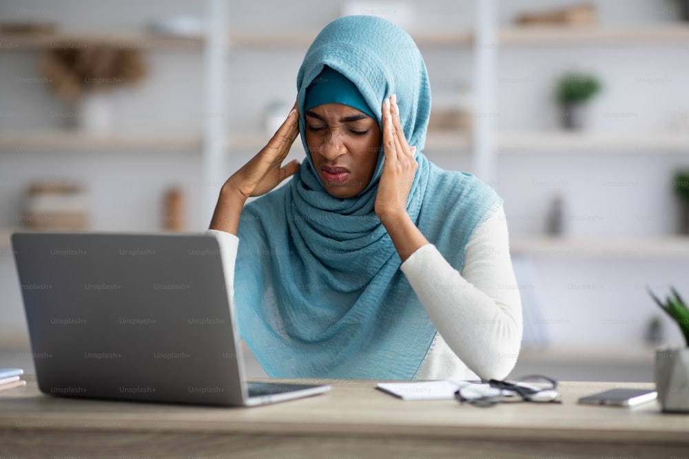 Headache Concept. Stressed Black Muslim Freelancer Lady Suffering From Migraine At Workplace, Young Annoyed African Islamic Woman Sitting At Desk With Laptop And Touching Temples, Free Space