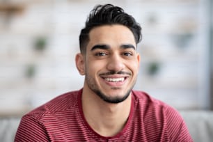 Portrait en gros plan d’un beau mec arabe souriant à la caméra tout en se relaxant à la maison, espace de copie. Jeune homme barbu du Moyen-Orient posant à la maison, vêtu d’une tenue décontractée