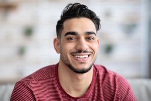 Retrato em close-up do alegre e bonito cara árabe sorrindo para a câmera enquanto relaxa em casa, espaço de cópia. Jovem positivo homem barbudo do Oriente Médio posando em casa, vestindo roupa casual