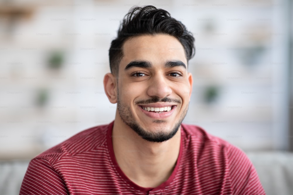 Closeup portrait of cheerful handsome arab guy smiling at camera while chilling at home, copy space. Positive young middle-eastern bearded man posing at home, wearing casual outfit