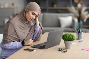 Trabajo estresante, concepto de agotamiento. Mujer musulmana estresada con pañuelo en la cabeza usando computadora portátil tocando la cabeza que tiene problemas en el lugar de trabajo sentada en la oficina en casa del escritorio. Crisis y problemas de negocios, concepto de dolor de cabeza