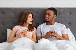 Vício em gadgets, phubbing, conceito de infidelidade matrimonial. Jovem casal inter-racial com smartphones sentados na cama, presos em gadgets, olhando um para o outro com desconfiança