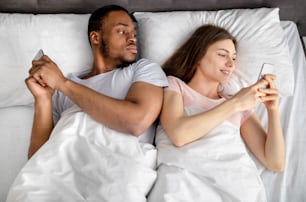 Jovem casal inter-racial deitado na cama preso em smartphones, jogando jogos online, navegando nas mídias sociais, vista superior. Problemas de relacionamento, vício em gadgets, infidelidade familiar, conceito de ciúme