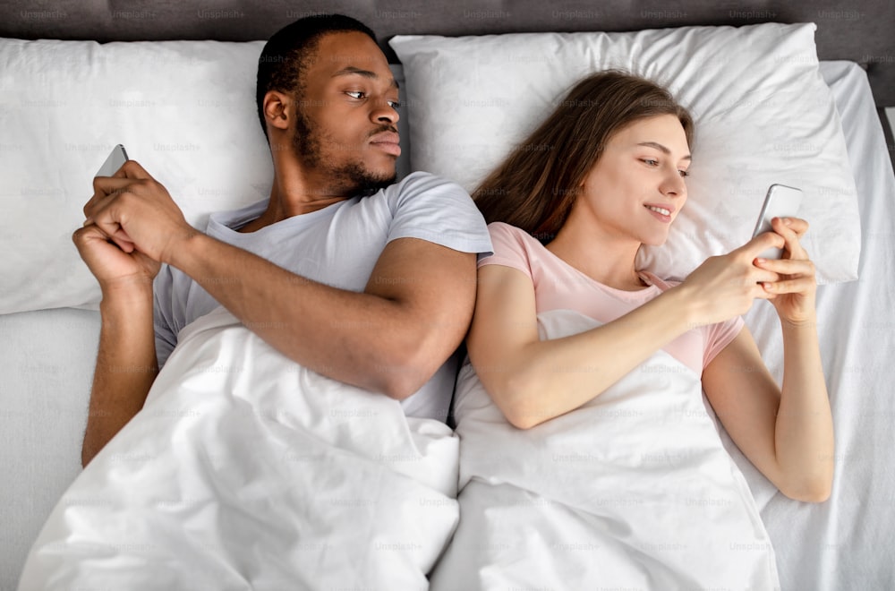 Young interracial couple lying in bed stuck in smartphones, playing online games, browsing social media, top view. Relationship problems, gadget addiction, family infidelity, jealousy concept