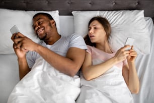 스마트폰 중독과 결혼 문제. 침대에서 모바일 기기를 사용하는 젊은 인종 간 커플, 서로에게 주의를 기울이지 않고, 온라인 게임을 하고, 웹 서핑을 하고, 평면도를 보인다