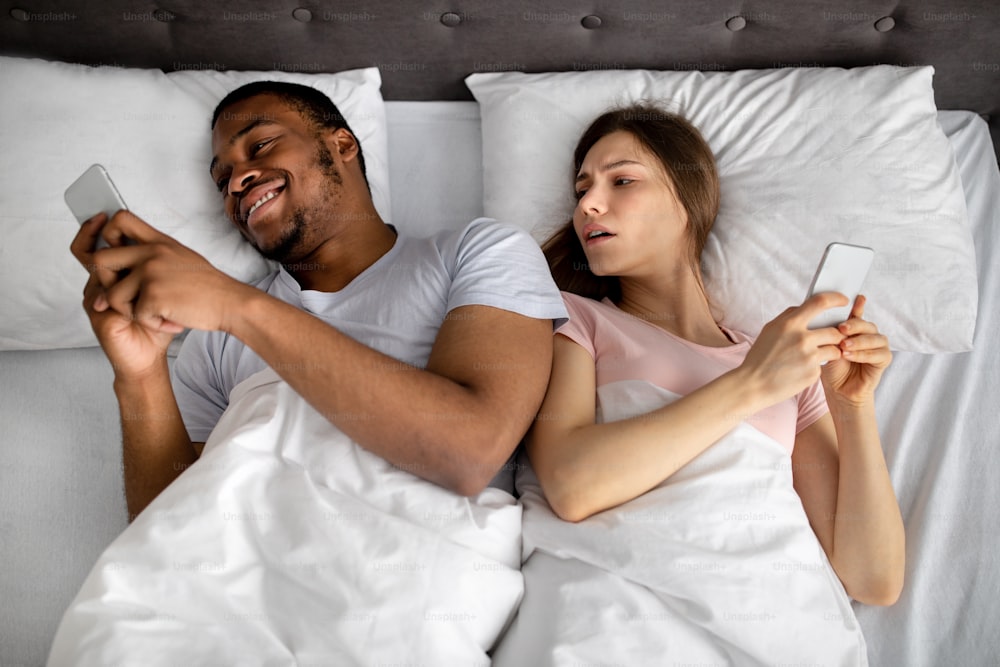 Smartphone addiction and marriage problems. Young interracial couple using mobile devices in bed, not paying attention to each other, playing online games, surfing web, top view