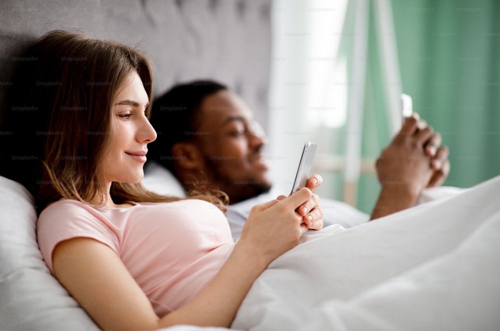 Gadget addiction concept. Young multiethnic family using smartphones while lying in bed. Caucasian wife and her black husband surfing internet or playing games, reading social media posts, copy space