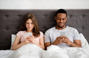 ガジェット中毒とファビング。携帯電話を持つ若い多国籍夫婦がベッドに座り、お互いを無視し、オンラインゲームやソーシャルネットワークで立ち往生し、インターネットを閲覧する