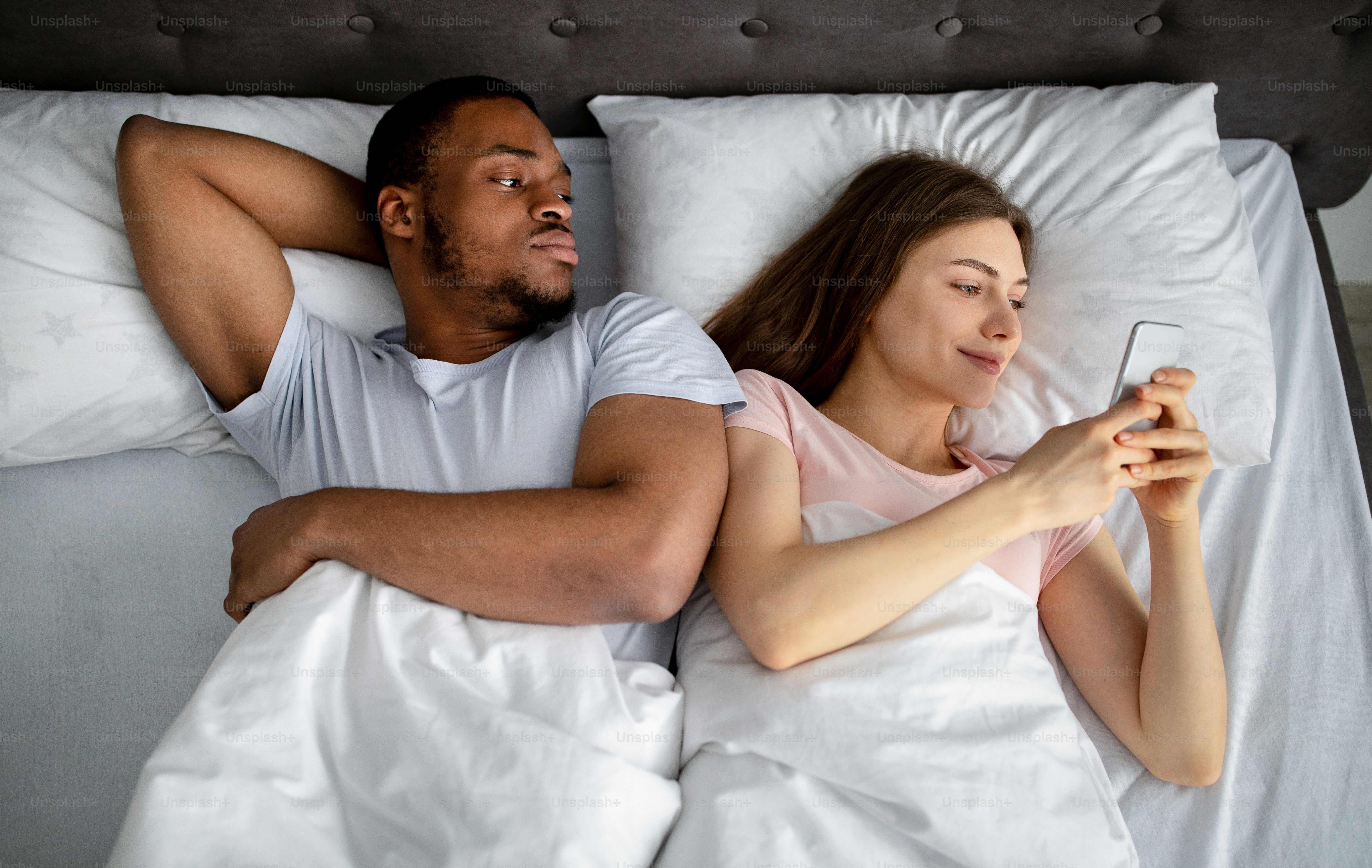 Irritated black guy feeling jealous of his girlfriend stuck in smartphone in bed, top view. Millennial man being neglected and ignored by his gadget addicted wife. Phubbing concept