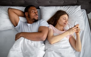 Irritated black guy feeling jealous of his girlfriend stuck in smartphone in bed, top view. Millennial man being neglected and ignored by his gadget addicted wife. Phubbing concept