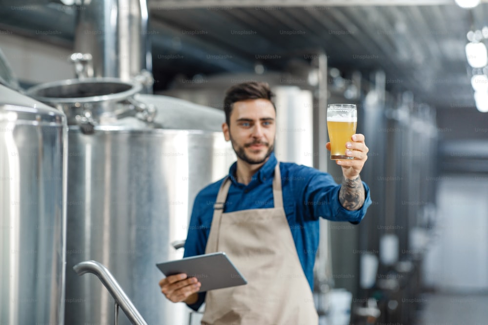 Male inspector working in distillery checks beer with app and gadget. Smiling millennial attractive man worker in apron, looks at glass of light craft drink, near kettles in brewery interior